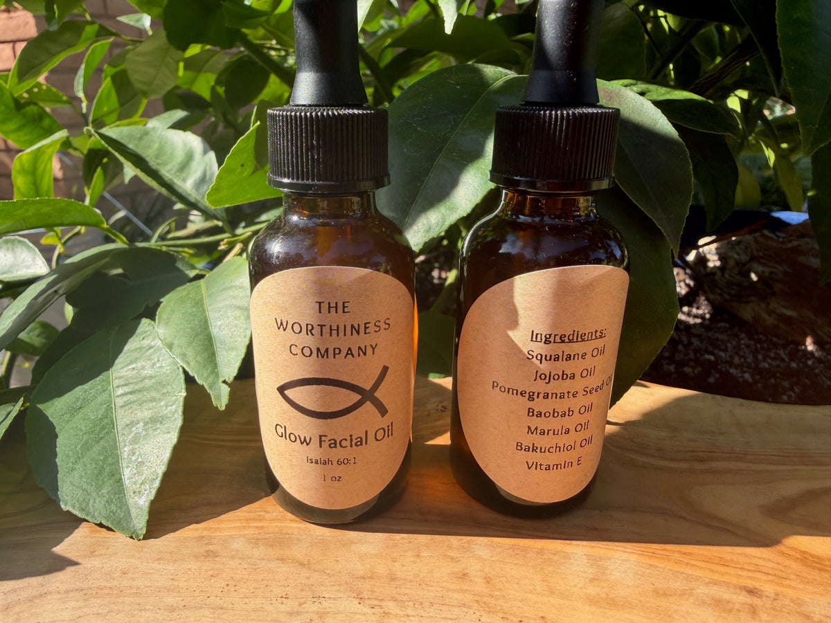 Two amber glass dropper bottles of facial oil with beige labels, placed on a wooden surface in front of green leaves. The left label reads 'THE WORTHINESS COMPANY Glow Facial Oil,' and the right bottle shows ingredient text including Squalane Oil, Jojoba Oil, Pomegranate Seed, Baobab Oil, Marula Oil, Bakuchiol Oil, and Vitamins E.