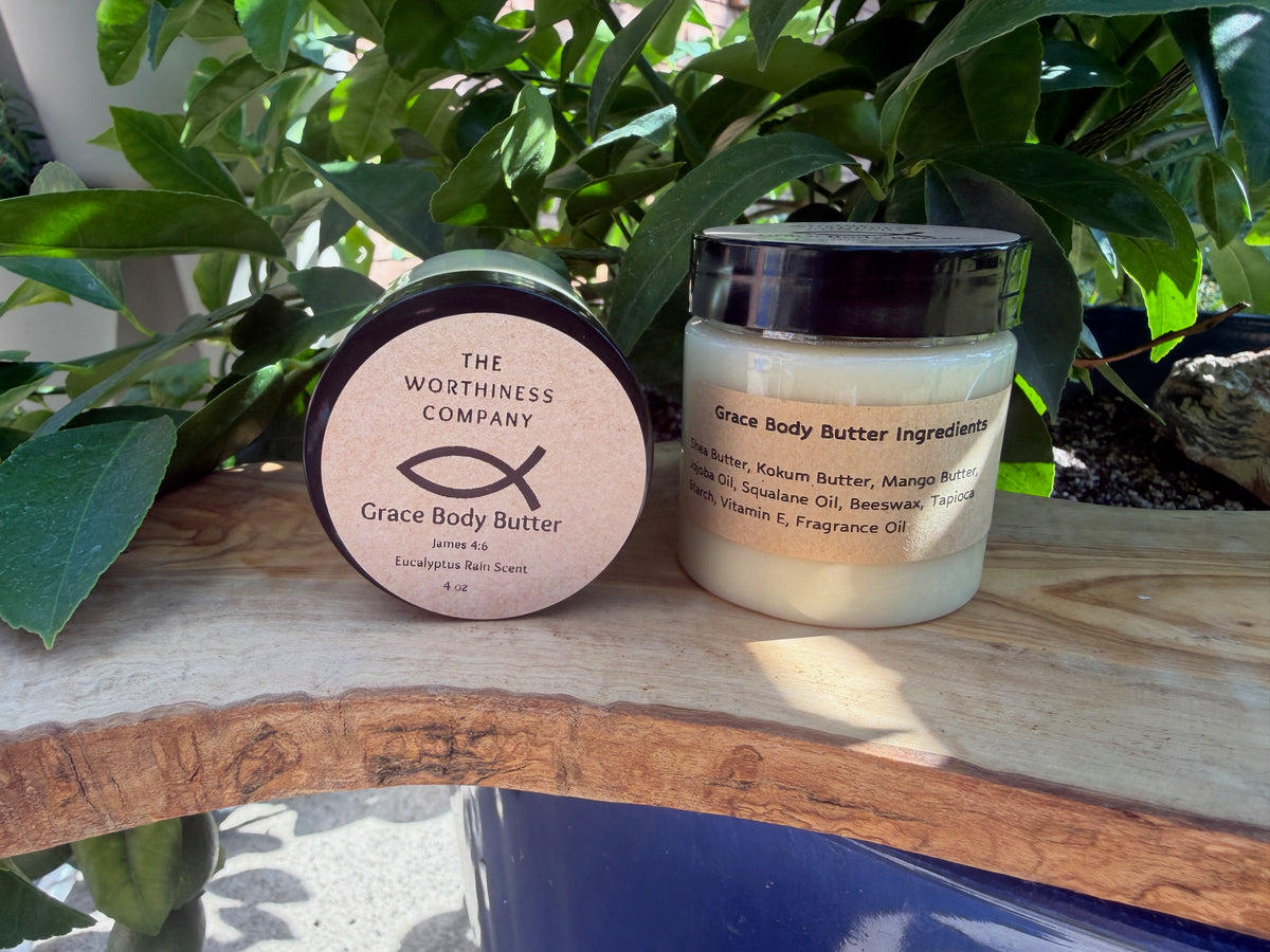 Two jars of The Worthiness Company Grace Body Butter placed on a wooden surface with green foliage in the background. 