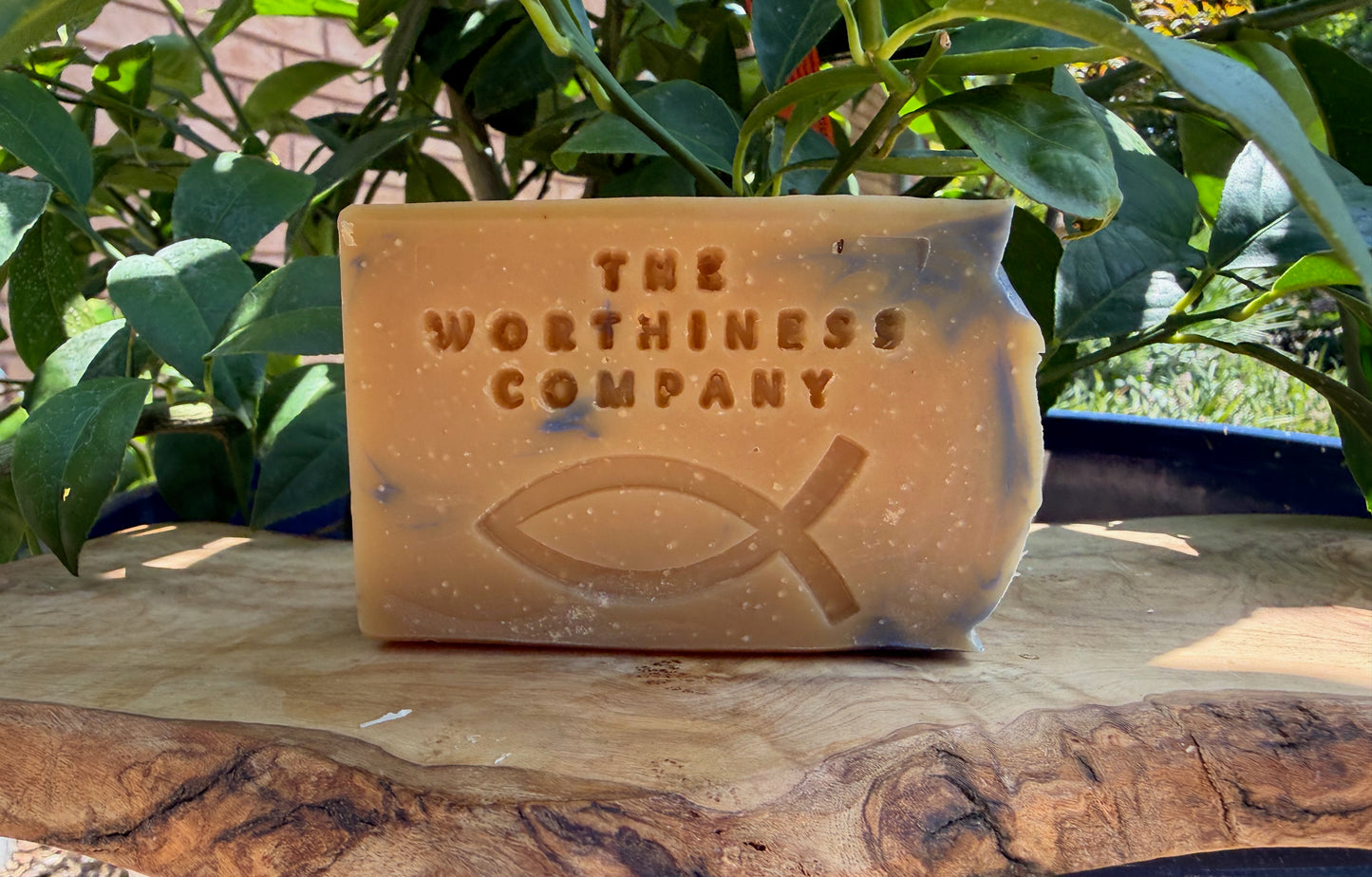 One andmade soap bar from The Worthiness Company displayed on a rustic wood slab.