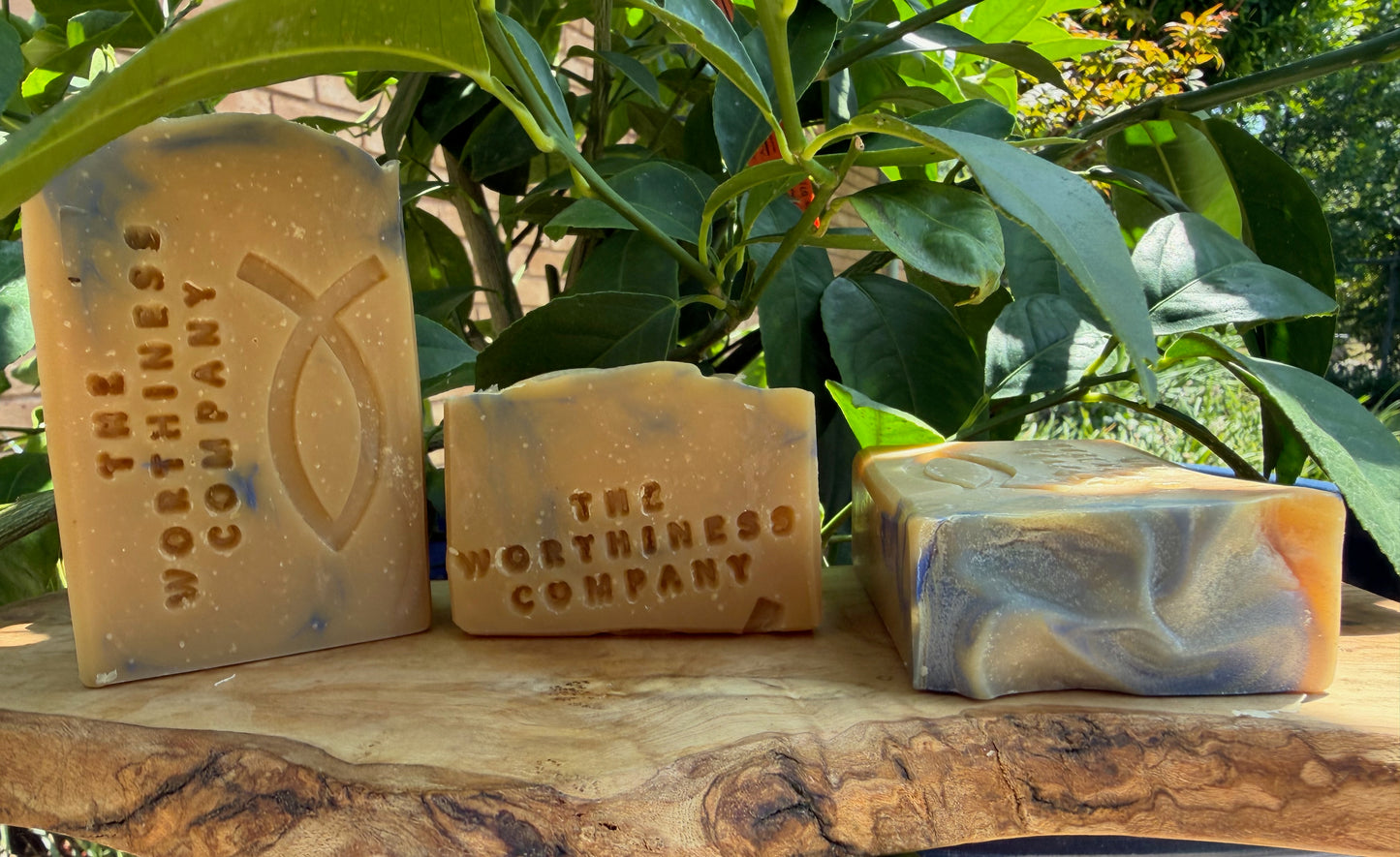 Three handmade soap bars from The Worthiness Company displayed on a rustic wood slab.