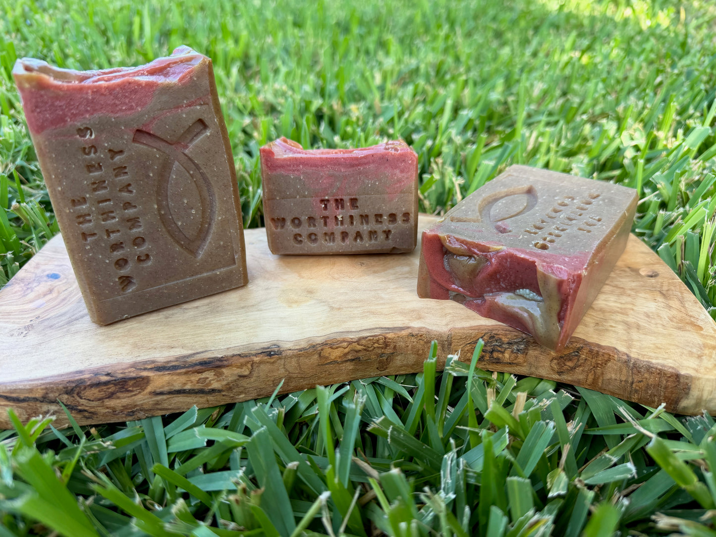 Three handmade soap bars from The Worthiness Company are displayed on a rustic wooden board set against. The soaps feature a distinct color story of dusty rose pink and earthy brown, with one bar showing a marbled swirl of both colors.