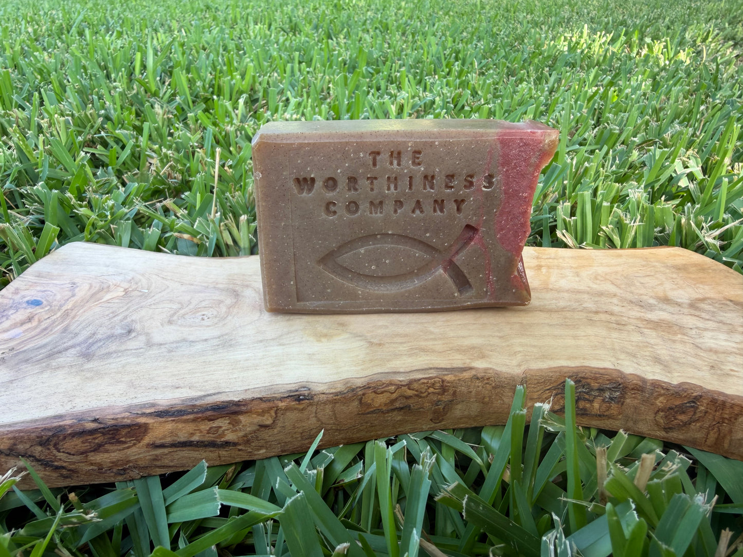 A rectangular brown soap bar embossed with 'THE WORTHINESS COMPANY' and a Christian fish symbol, resting on a wooden board.