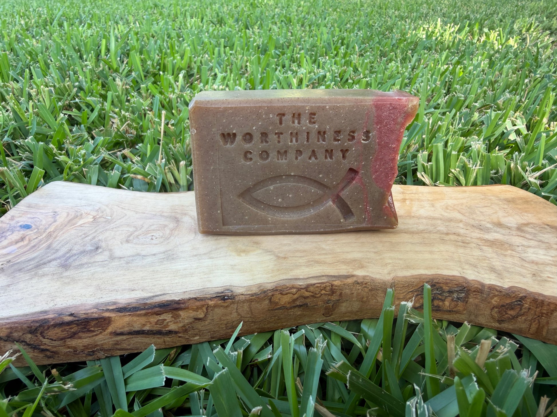 A rectangular brown soap bar embossed with 'THE WORTHINESS COMPANY' and a Christian fish symbol, resting on a wooden board.