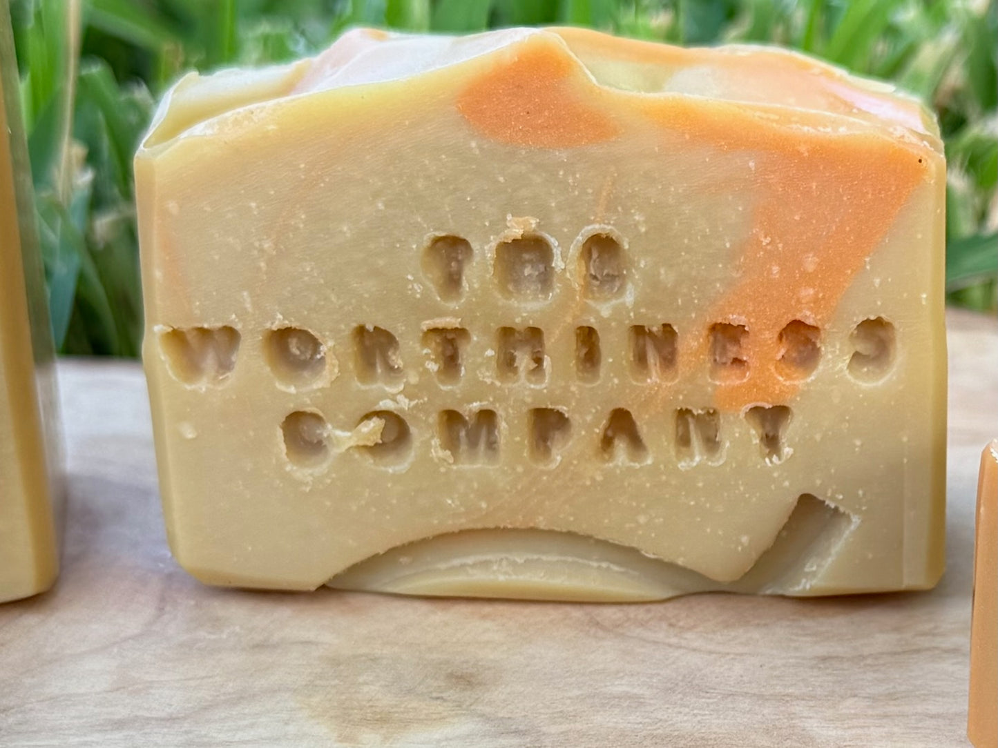 A half-bar  handmade soap bar stamped with 'The Worthiness Company' on the front, featuring a creamy beige base with orange swirls, resting on a wooden surface with green foliage in the background.
