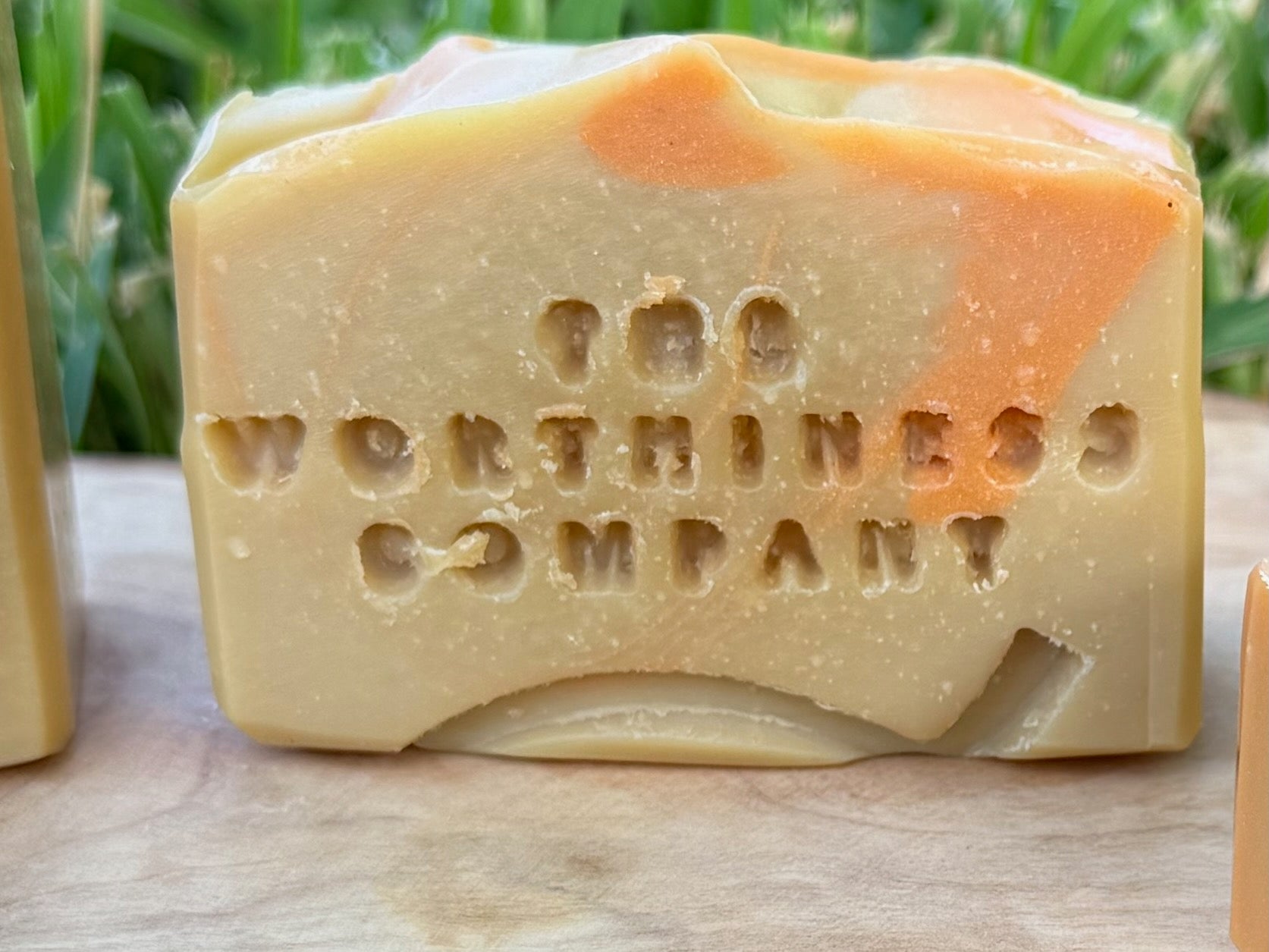 A half-bar  handmade soap bar stamped with 'The Worthiness Company' on the front, featuring a creamy beige base with orange swirls, resting on a wooden surface with green foliage in the background.