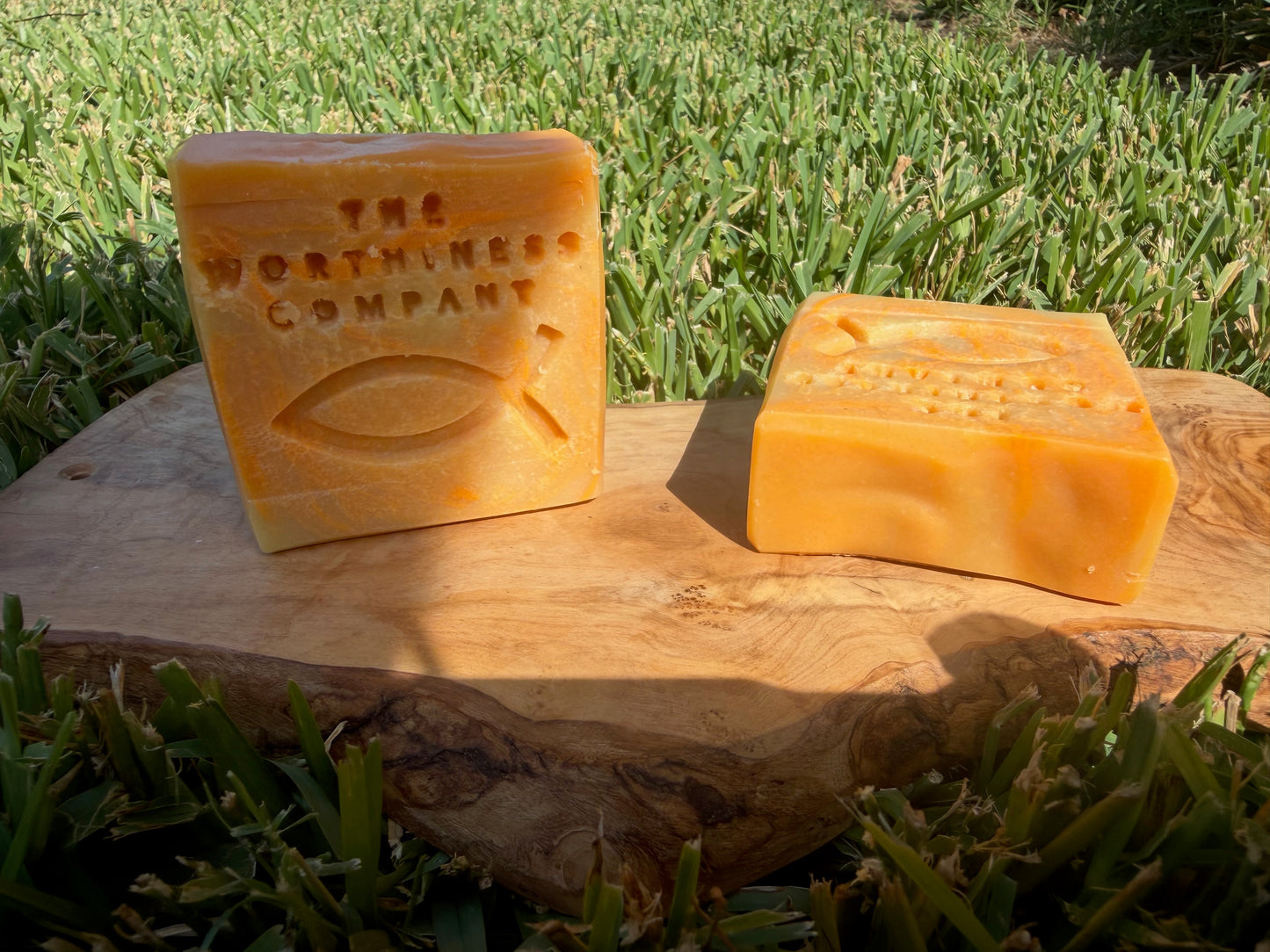 Two rectangular bars of orange-yellow soap displayed on a natural wood slab outdoors, with green grass in the background. One bar is embossed with the text 'THE WORTHINES COMPANY' and the christian fish symbol.
