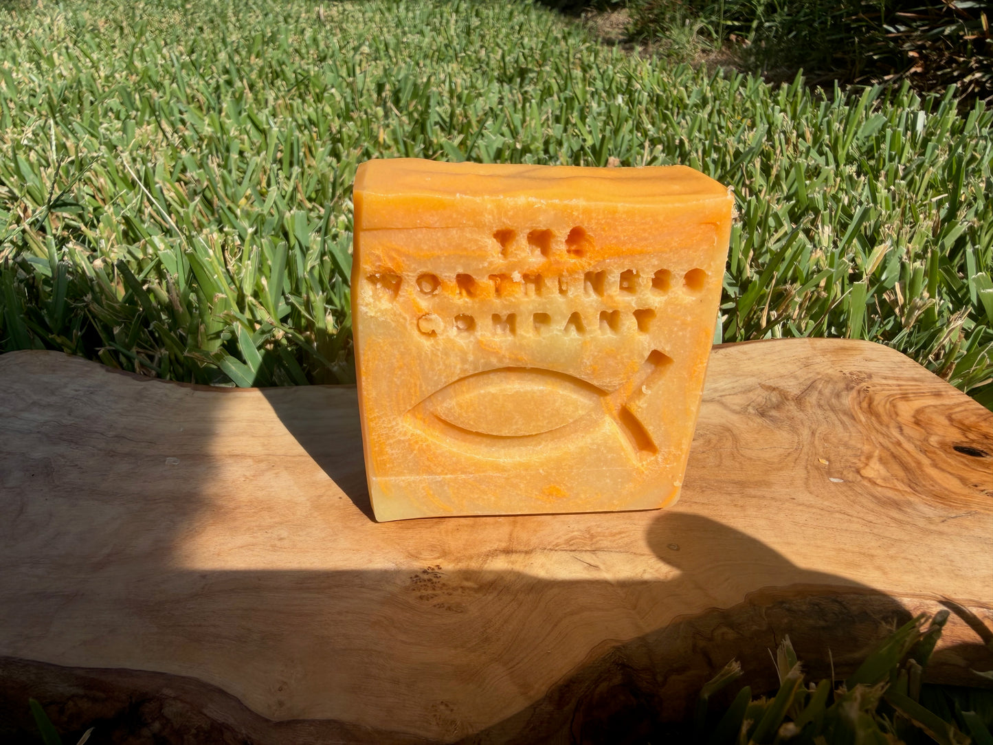 An orange-yellow rectangular bar of soap standing upright on a natural wood slab outdoors. The soap is embossed with the text 'THE WORTHINESS COMPANY' and a christian fish symbol.
