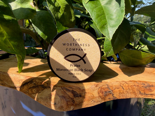 A round container of The Worthiness Company Praise Moroccan Black Soap sits on a rustic live-edge wooden slab.