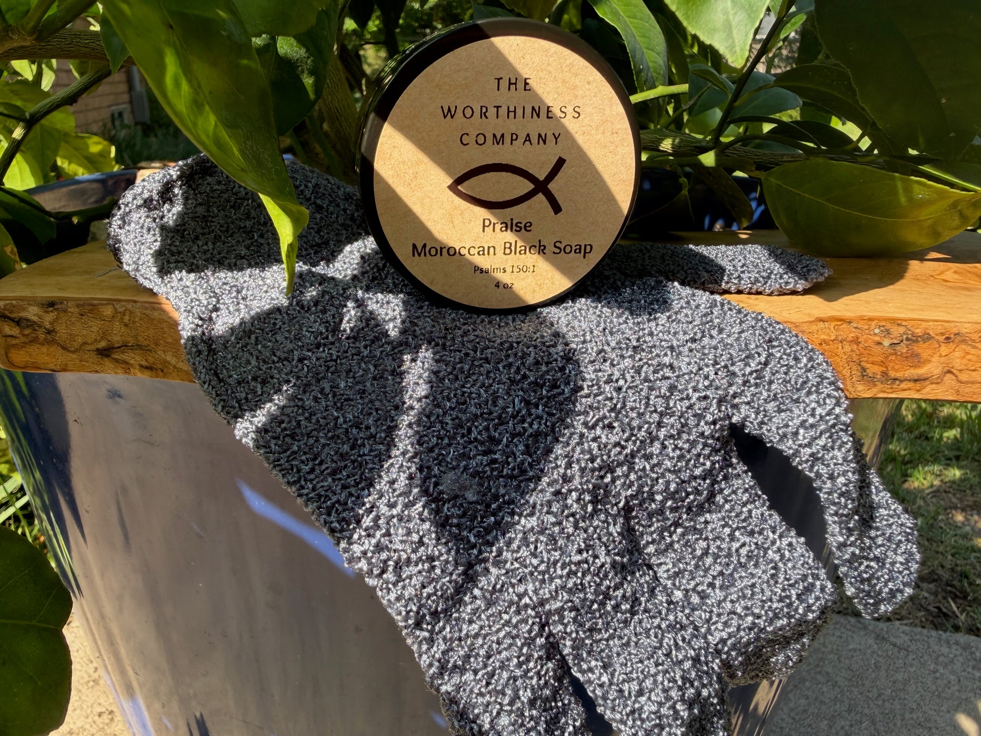 A round container of The Worthiness Company 'Praise Moroccan Black Soap' on a wooden surface draped with an exfoliating glove.