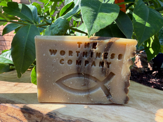 A rectangular bar of handmade soap from 'The Worthiness Company' with an embossed Ichthys symbol, resting on a wooden surface with green leaves in the background.