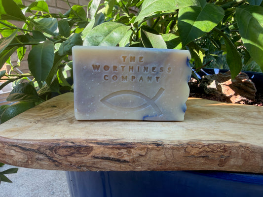 A rectangular bar of soap from The Worthiness Company sits on a rustic wooden board outdoors, surrounded by green leaves and natural sunlight.