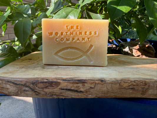 A rectangular bar of soap in a warm peach-beige color, embossed with the text 'THE WORTHINESE COMPANY' and a Christian fish symbol, resting on a rustic wooden board with green leaves in the background.