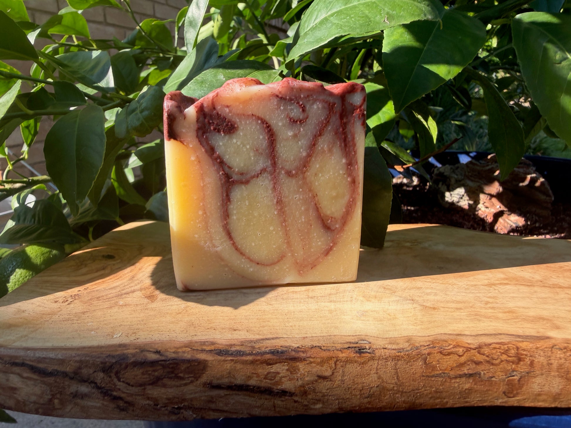 A rectangular bar of handmade soap with a marbled pattern of cream, yellow, and reddish-brown, standing on a rustic wooden board with green leaves in the background.