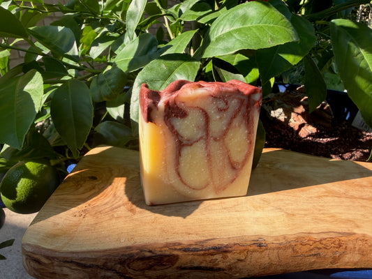 A rectangular bar of handmade soap with a marbled pattern of pale yellow, cream, and reddish-brown, displayed on a rustic wooden board with green citrus leaves in the background.