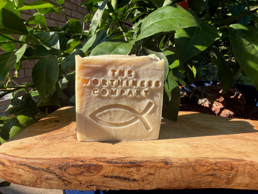 A rectangular bar of soap from The Worthiness Company, embossed with a Christian fish symbol and brand name, standing on a rustic wooden slab with green leaves in the background.