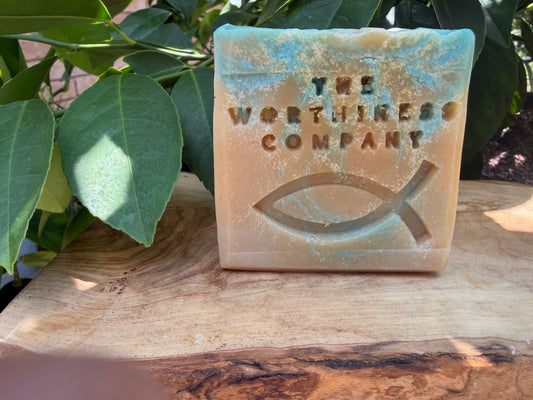 A rectangular bar of soap from The Worthiness Company sits on a wooden surface outdoors, with green leaves in the background. The soap has a warm peach-beige base with turquoise-blue marbling and a cutout fish symbol on the front.