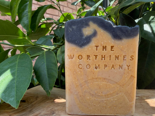 A rectangular bar of soap with a rustic, irregular top edge, featuring a speckled tan and orange body with a dark charcoal-black swirled top section. The soap is embossed with the text 'THE WORTHINESS COMPANY'.