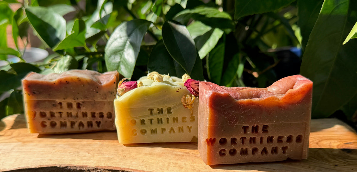 Three half bars of soap from The Worthiness Company displayed on a wooden board with green foliage in the background. The soap bars feature earthy brown, pale yellow with dried flower accents, and terracotta orange-red tones.