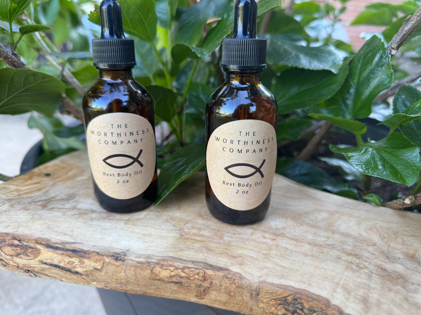 Two amber glass dropper bottles of The Worthiness Company  Body Oil resting on a rustic wood slice.