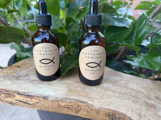 Two amber glass dropper bottles of The Worthiness Company  Body Oil resting on a rustic wood slice.