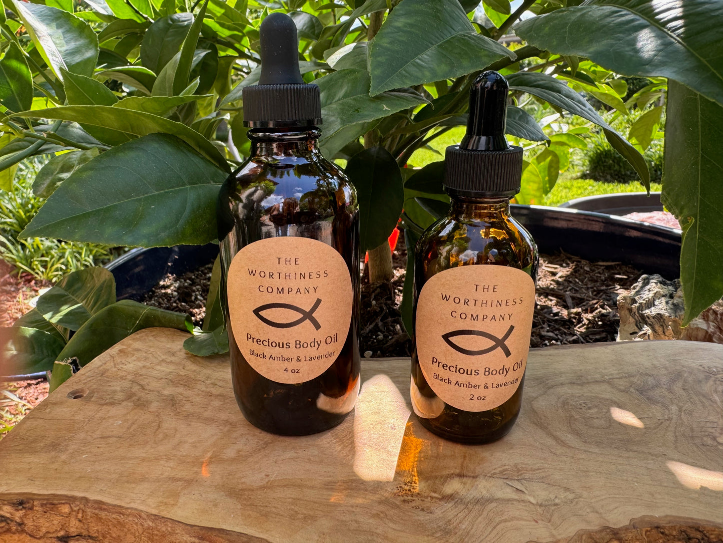 Two amber glass dropper bottles of The Worthiness Body Oil with black caps and beige labels, placed on a wooden surface.