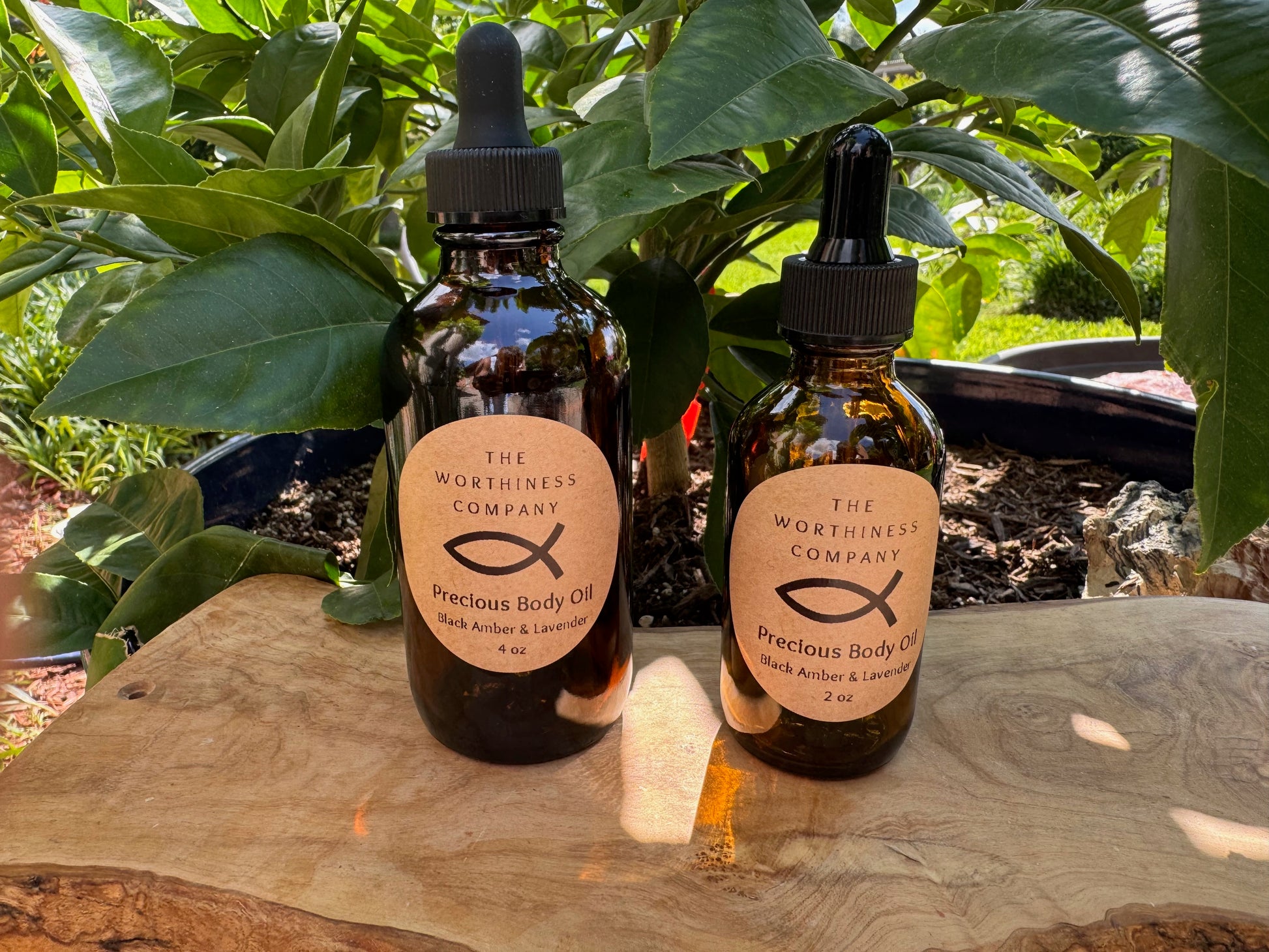 Two amber glass dropper bottles of The Worthiness Body Oil with black caps and beige labels, placed on a wooden surface.