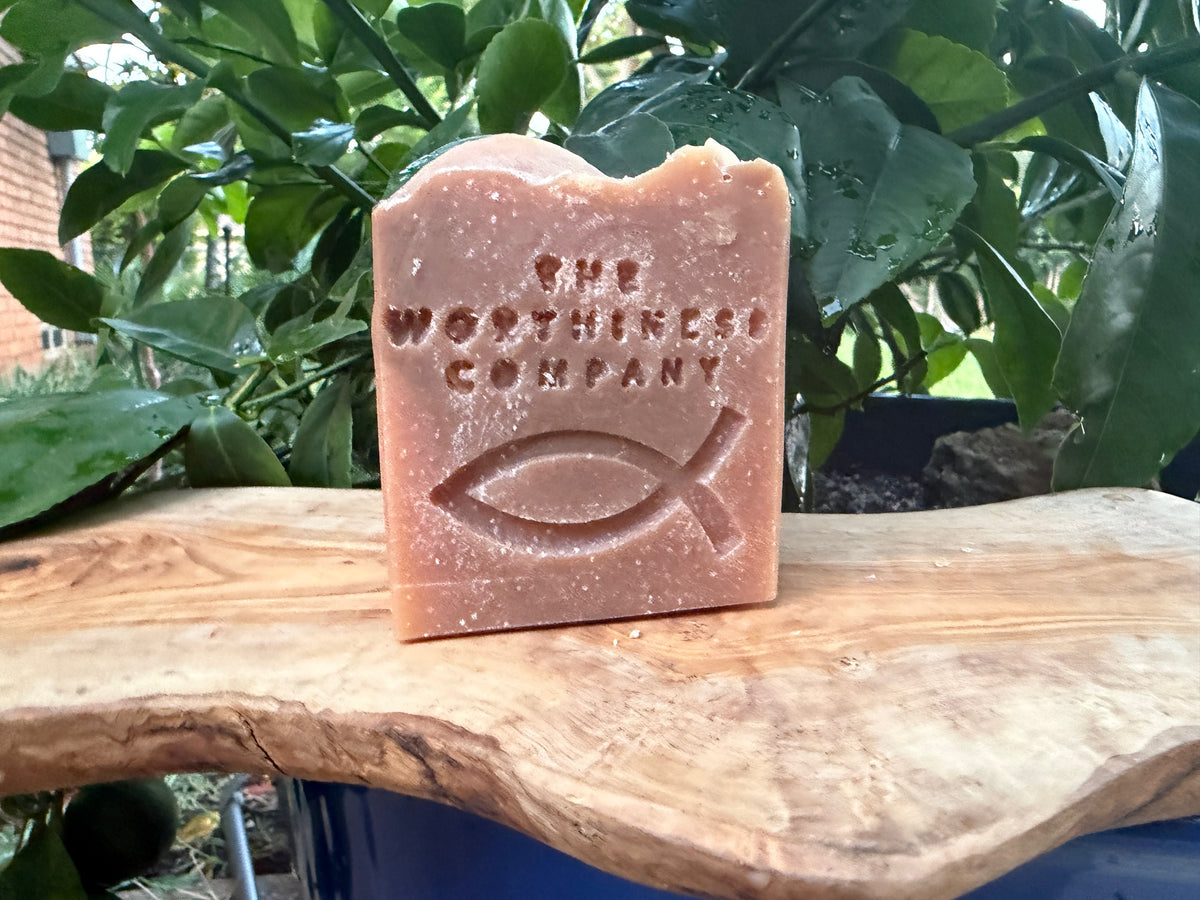 A rectangular bar of soap with a rustic, hand-broken top edge, embossed with the text 'THE WORTHINESS COMPANY' and a Christian fish symbol. 