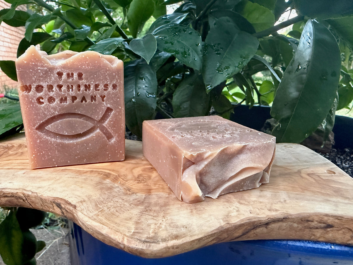 Two handmade soap bars in a natural outdoor setting on a wooden slab with green plants in the background. One bar is embossed with 'THE WORTHINESS COMPANY' and a Christian ish symbol,