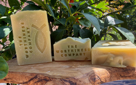 Three bars of handmade soap from The Worthiness Company displayed on a rustic wood slab.