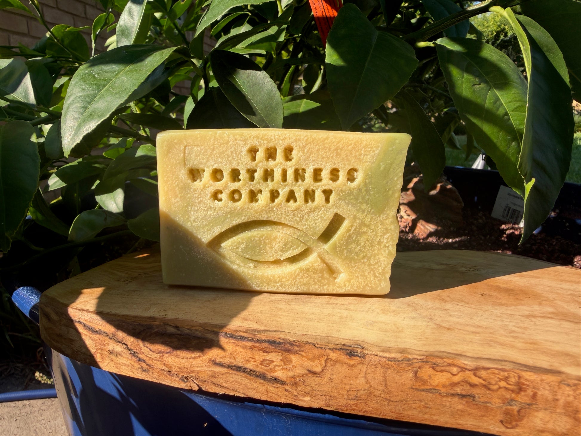 A rectangular yellow stone plaque with the text 'THE WORTHINESS COMPANY' and an ichthys symbol, displayed on a wooden surface with green foliage in the background.